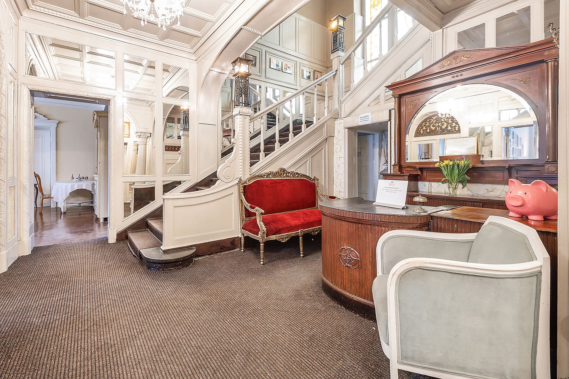 Elegancki hotelowy hol z białą parterową balustradą, czerwonym welurowym kanapą i przeszklonymi drzwiami; od lewej widok na małą klapę/drzwi i stół z białą tkaniną w oddali, na pierwszym planie elegancki drewniany całkowicie wyściełany blat recepcji z dużym lustrem i różowym granatowym kolorem.