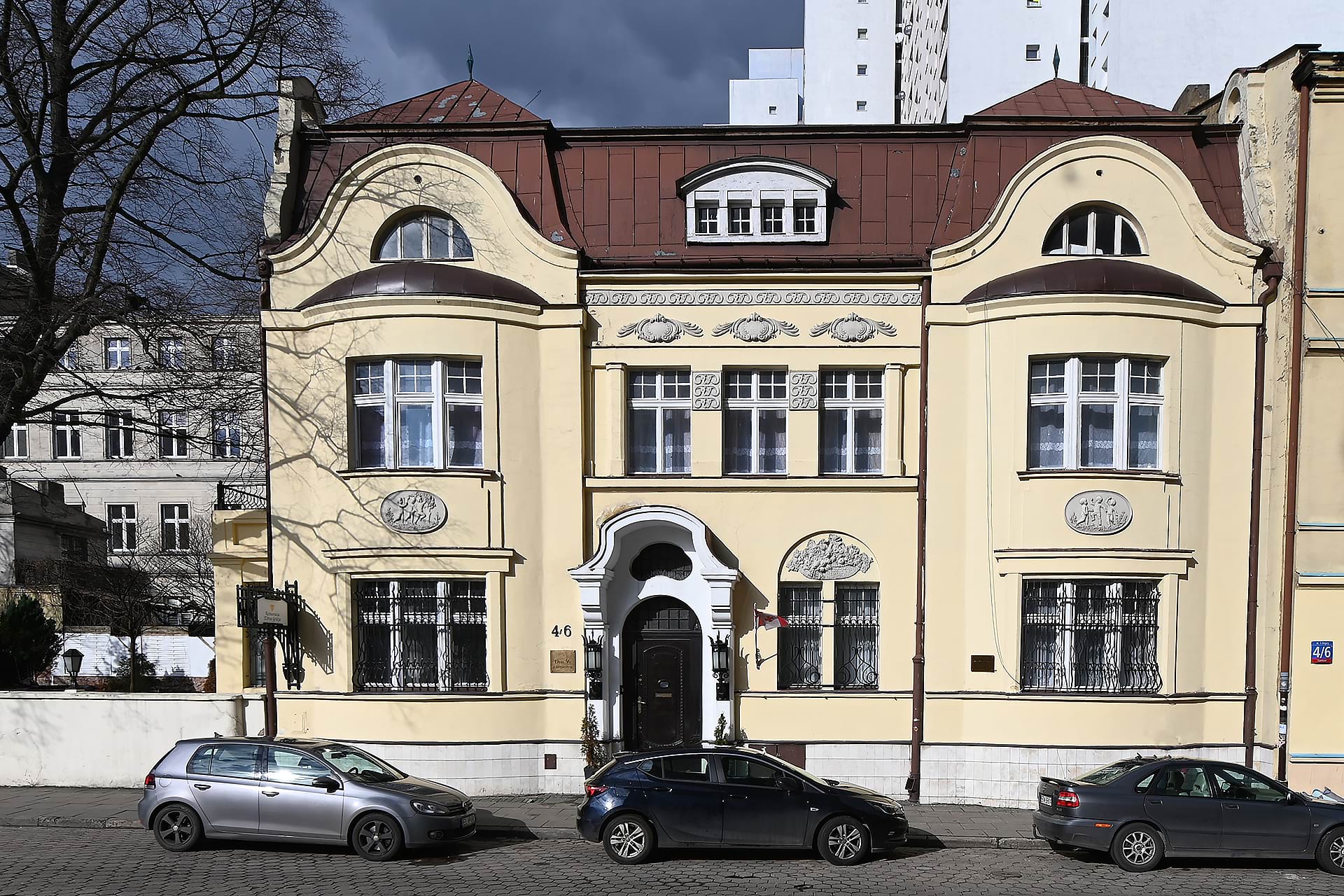 Żółta kamienica o zdobionej fasadzie, dwie dużej szerokości okna, centralne wejście z łukową podeszwą i trójkątnymi zdobieniami, zaparkowane samochody na ulicy