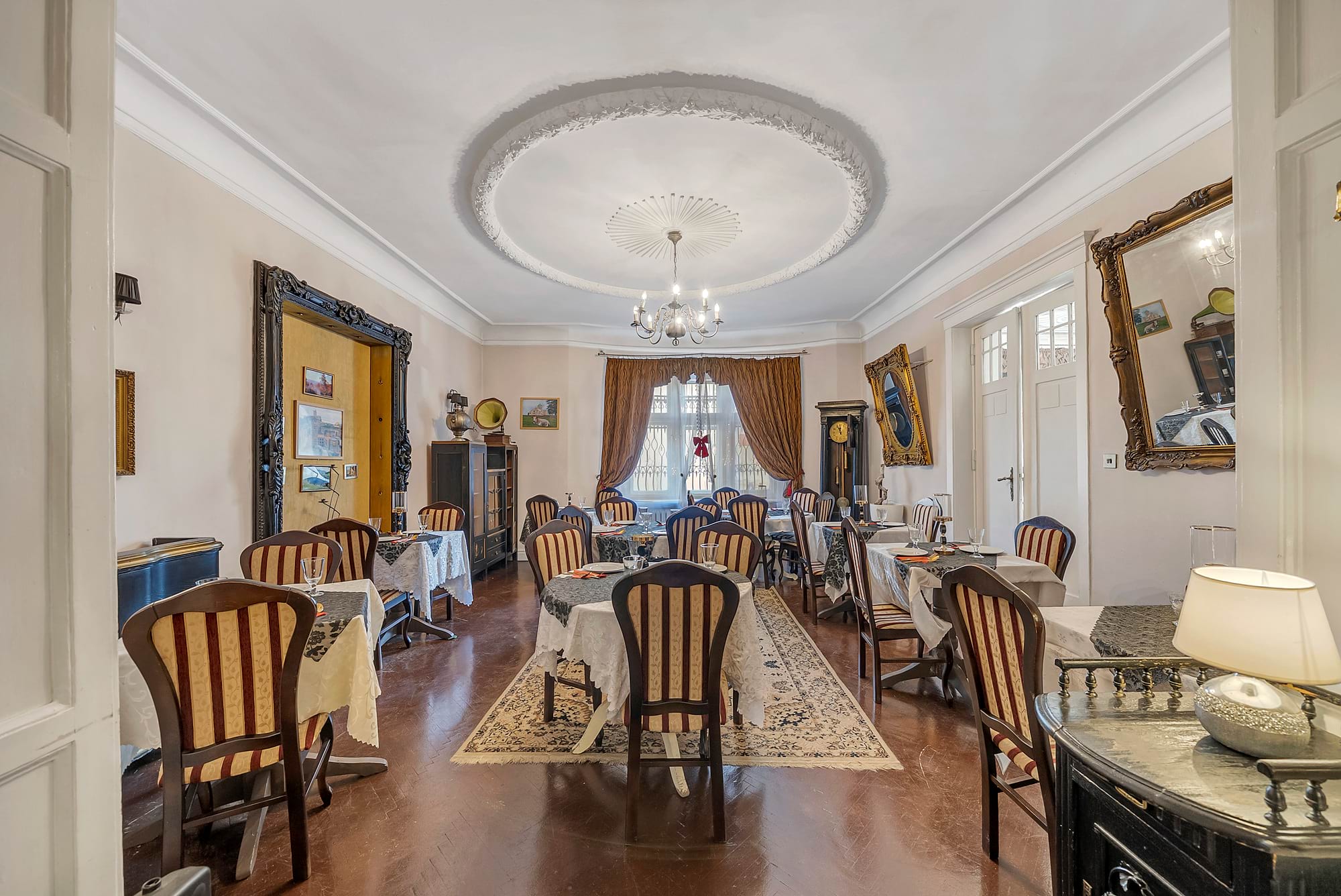 Elegancka jadalnia w klasycznym stylu z okazałym sufitem, centralnym żyrandolem i oknami z zasłonami; stoliki nakryte białymi obrusami, krzesła w czerwono-paskowanej tapicerce, antyczne meble i obrazy na ścianach