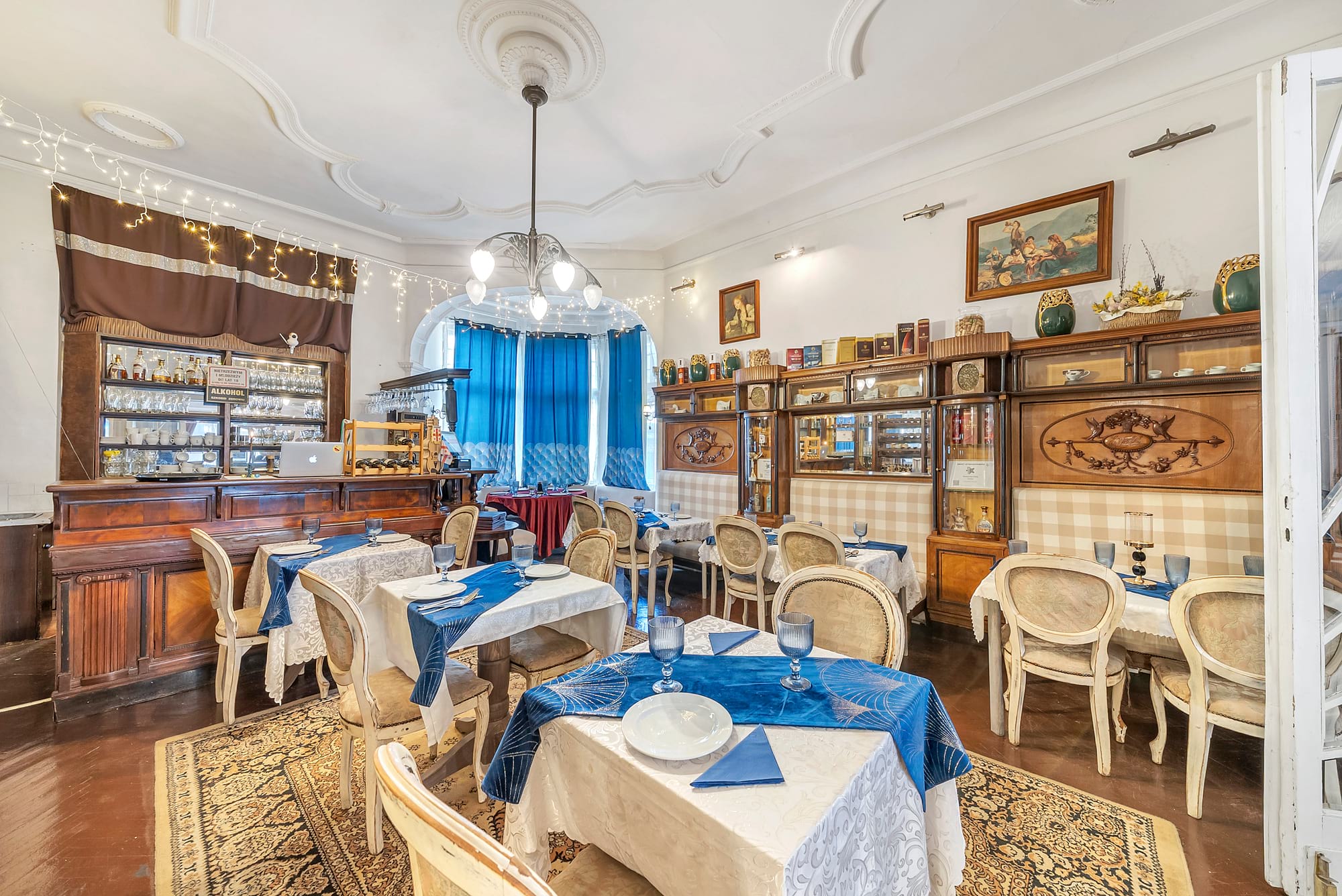 Elegancka jadalnia w stylu retro z drewnianymi meblami, błękitnymi zasłonami i stołami z białymi obrusami oraz niebieskimi dodatkami